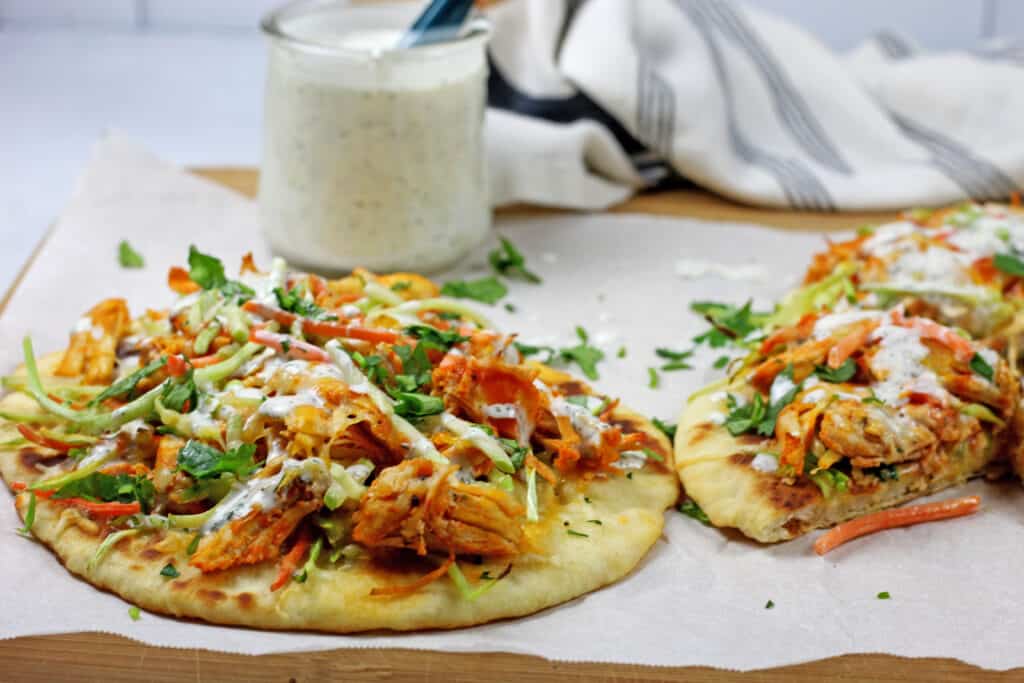 Buffalo chicken flatbread on parchment paper with a cup of dill sauce with a blue spoon in it is in the background. There is also a blue and white napkin in the background.