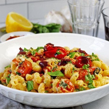 Picture of the tiktok feta pasta with cherry tomatoes in a white bowl with lemon, basil, and wine glasses in the background. One of the wine glasses has a red wine in it.