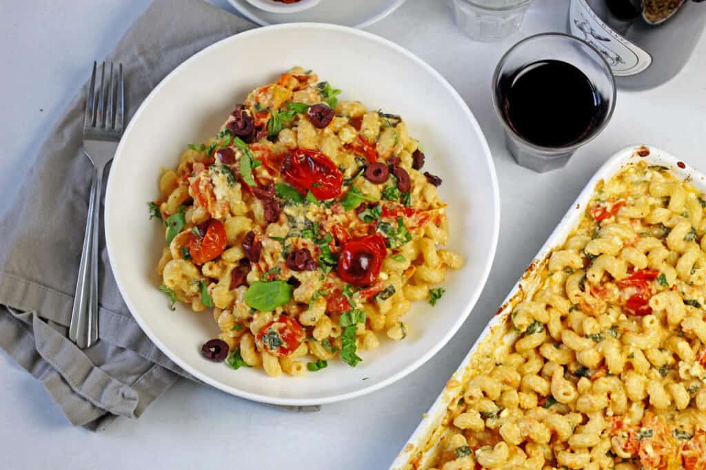 Overview of tiktok feta pasta with cherry tomatoes in a white bowl on a white counter. On the side is a white baking dish with more pasta, a glass of red wine, and a grey napkin with a fork.