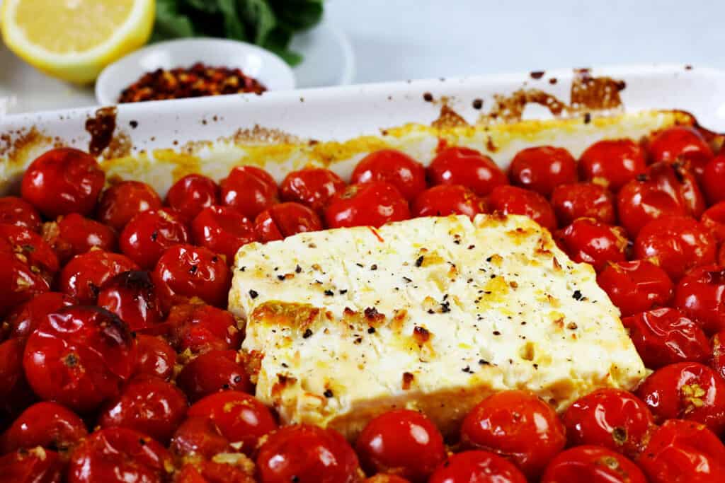 A close up of the feta with the cherry tomatoes baked in a white baking dish. The tops of the tomatoes and feta cheese are browned and blistered. There is lemon, basil, and red pepper flake in the background.