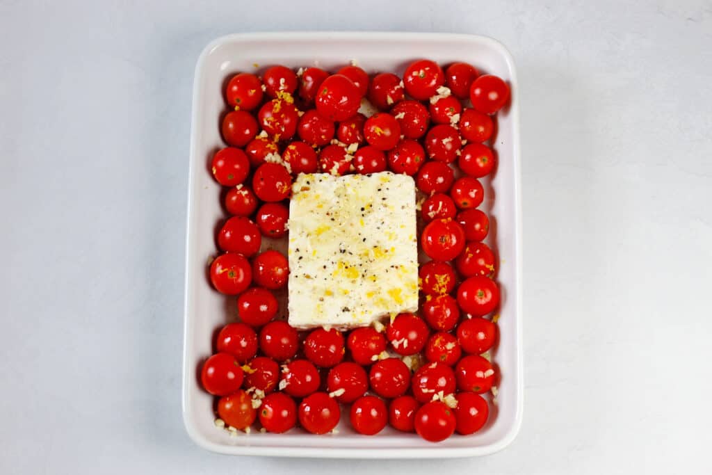 An overhead shot of the feta cheese block in the middle of the cherry tomatoes in a white baking dish on a white countertop. Seasonings and lemon zest are sprinkled on top of the feta cheese.