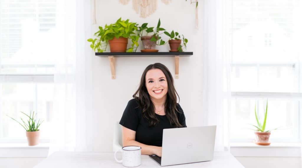 Melissa Macher in an office with white walls and desk working at a laptop. A cup of coffee is on the side of the laptop and there are plants in the background.