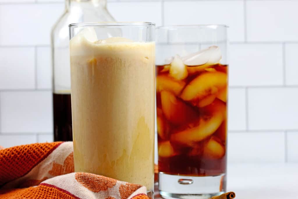 Two tall glasses of pumpkin cold brew sitting on an orange towel in front of white subway tile. One cold brew has cream in it and one is black. There is plain black cold brew behind the glasses of pumpkin cold brew.