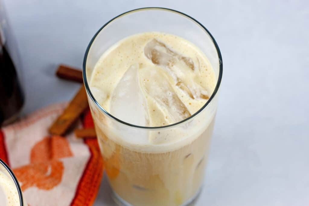 An overhead view of a glass of pumpkin cold brew with ice sitting next to an orange towel and cinnamon sticks on a white background.