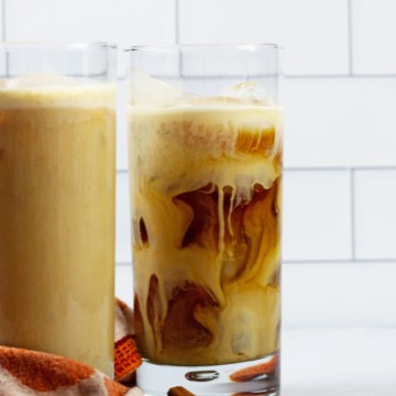 Two tall glasses of pumpkin cold brew sitting on an orange towel in front of white subway tile.