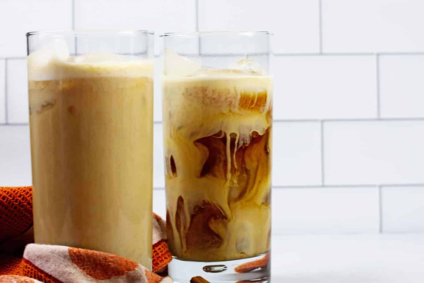 Two tall glasses of pumpkin cold brew sitting on an orange towel in front of white subway tile.