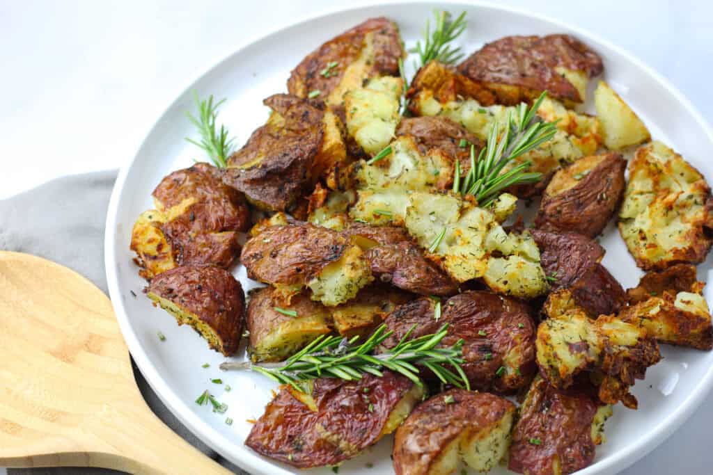 A plate of air fryer smashed potatoes on a white plate on a white countertop with a gray napkin and garnished with rosemary. A wooden spoon rests upon the gray napkin.