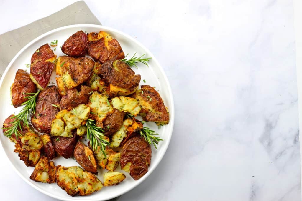 A plate of air fryer smashed potatoes on a white plate on a white countertop with a gray napkin and garnished with rosemary.