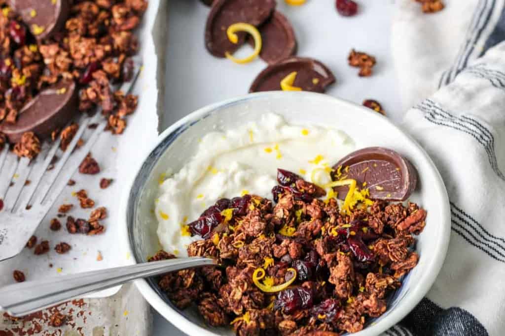 Bowl of Christmas granola with yogurt, orange zest, and chocolate orange slices with a sheet pan of Christmas granola in the background.