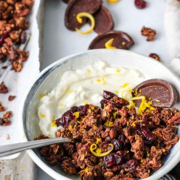 Bowl of Christmas granola with yogurt, orange zest, and chocolate orange slices with a sheet pan of Christmas granola in the background.