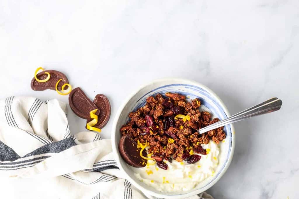 Bowl of Christmas granola and yogurt topped with chocolate and orange zest on white counter with a blue and white napkin, chocolate, and zest scattered about.