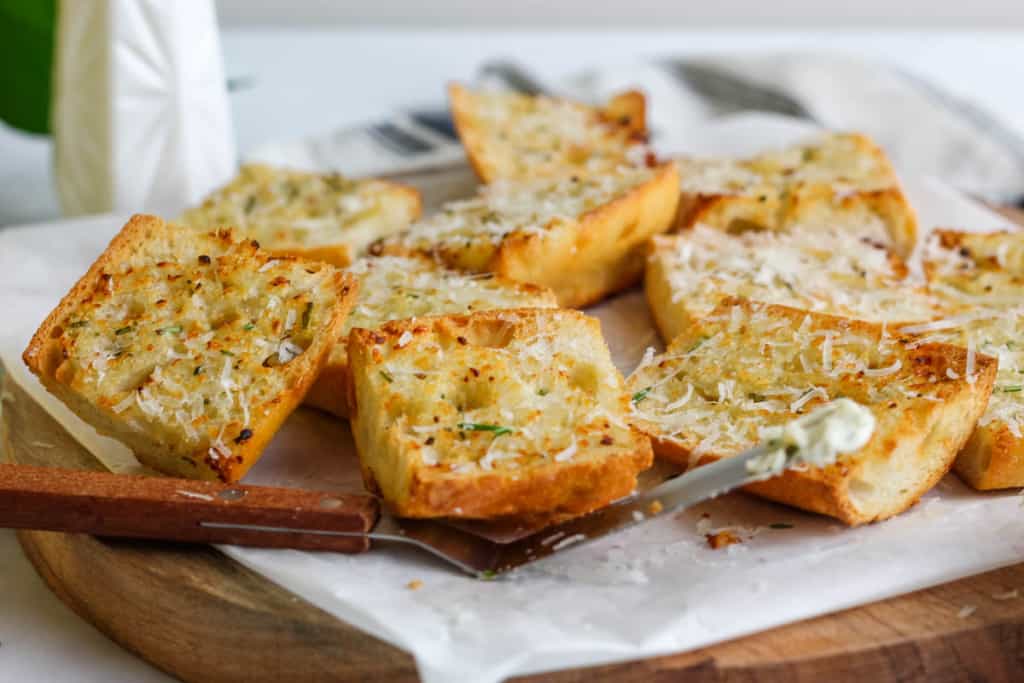 Slides of air fryer garlic bread with a butter knife on white paper on a wood cutting board.