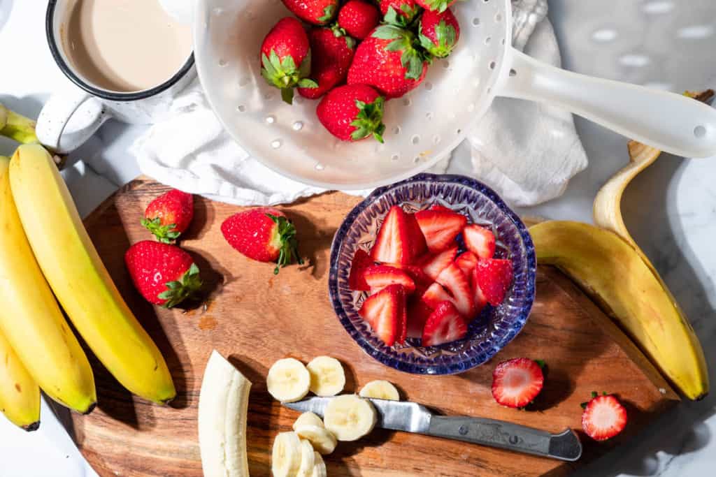 Overview of sliced banana and strawberries on a cutting board with a colander of strawberries on the side and a cup of coffee.