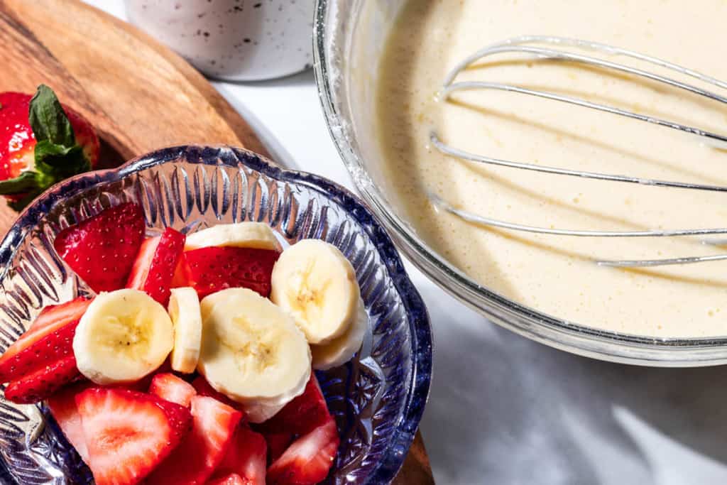 A blue bowl of sliced strawberries and bananas with a bowl of pancake batter and a whisk.