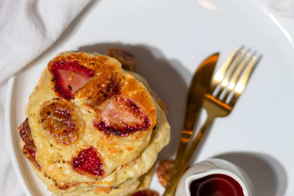 Top view of the strawberry banana pancakes studded with the fruit on a white plate with syrup and gold silverware on the side.