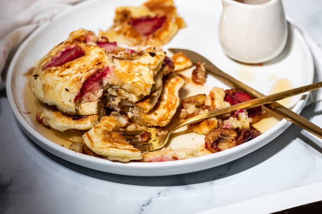 A messy plate of strawberry banana pancakes with bite size pieces scattered on a white plate with a white container of syrup and gold flatware.