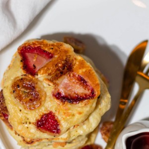 Top view of the strawberry banana pancakes studded with the fruit on a white plate with syrup and gold silverware on the side.