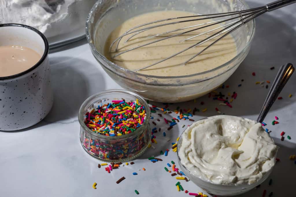 Bowl of pancake batter with a whisk sitting inside. Another bowl with rainbow sprinkles, and another bowl with whipped cream on a marble countertop. A cup of coffee sits off to the side.