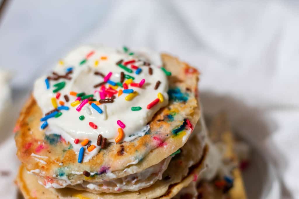 A stack of rainbow confetti pancakes with a dollop of whipped cream on top with extra sprinkles.