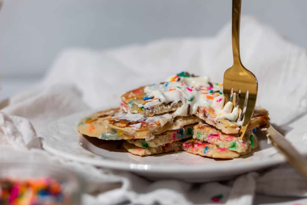 A stack of rainbow confetti sprinkle pancakes with whipped cream on top with a bite taken out of the middle with a gold fork.