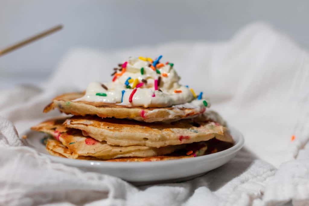 Stack of confetti pancakes with whipped cream on top on a white plate and white towel.