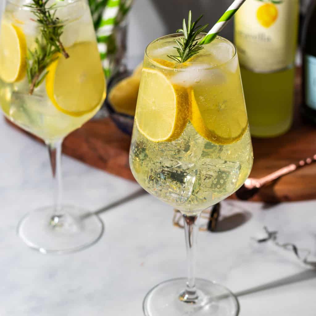 Two glasses of limoncello spritz with garnishes of lemons and rosemary. In the background a cutting board with limoncello and straws sits.