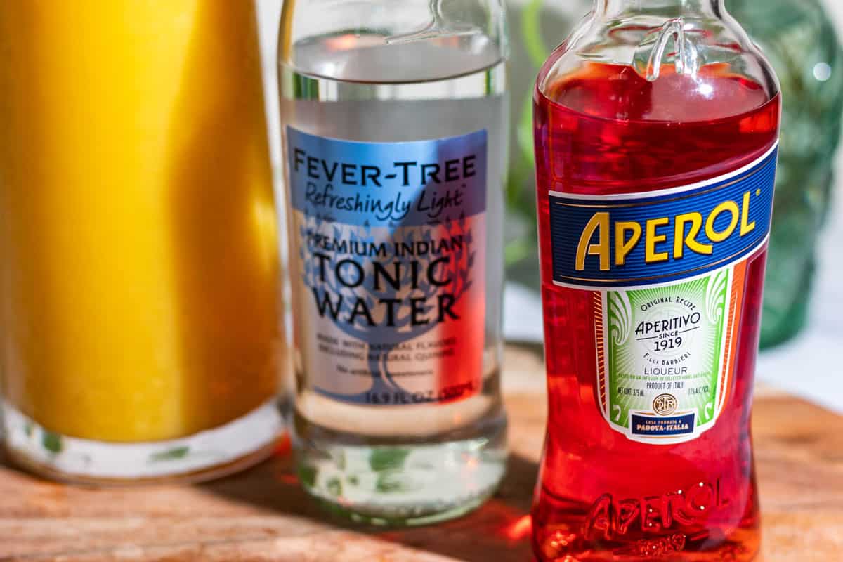 Ingredients for the aperol tonic cocktail with an aperol bottle, tonic water bottle, and a carafe of orange juice.