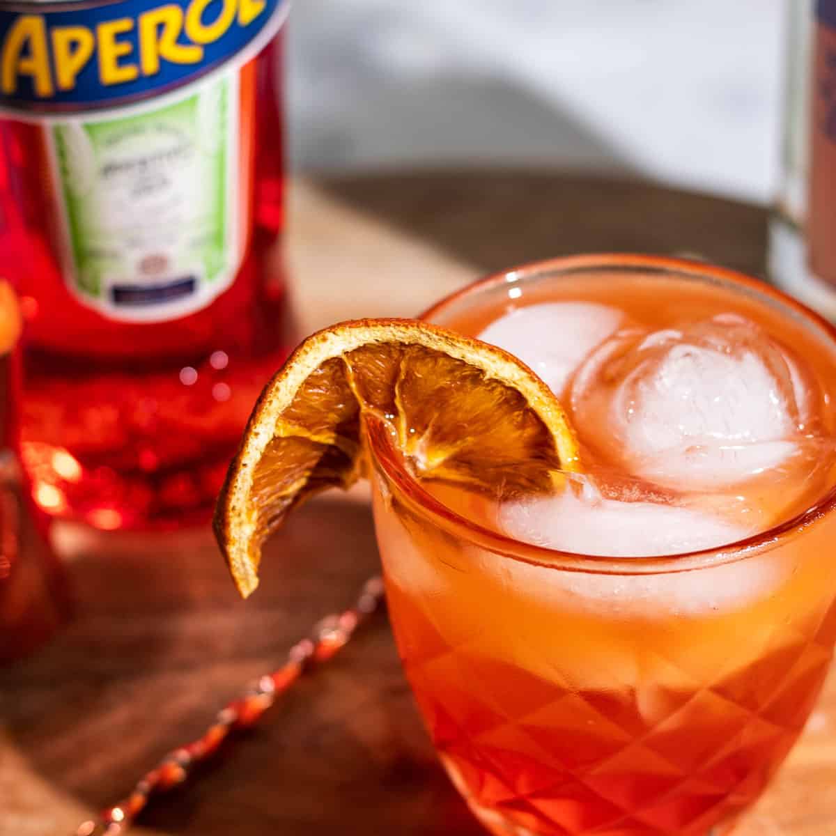 Glass of aperol tonic on a wood surface with a cocktail spoon and a bottle of aperol on the side.