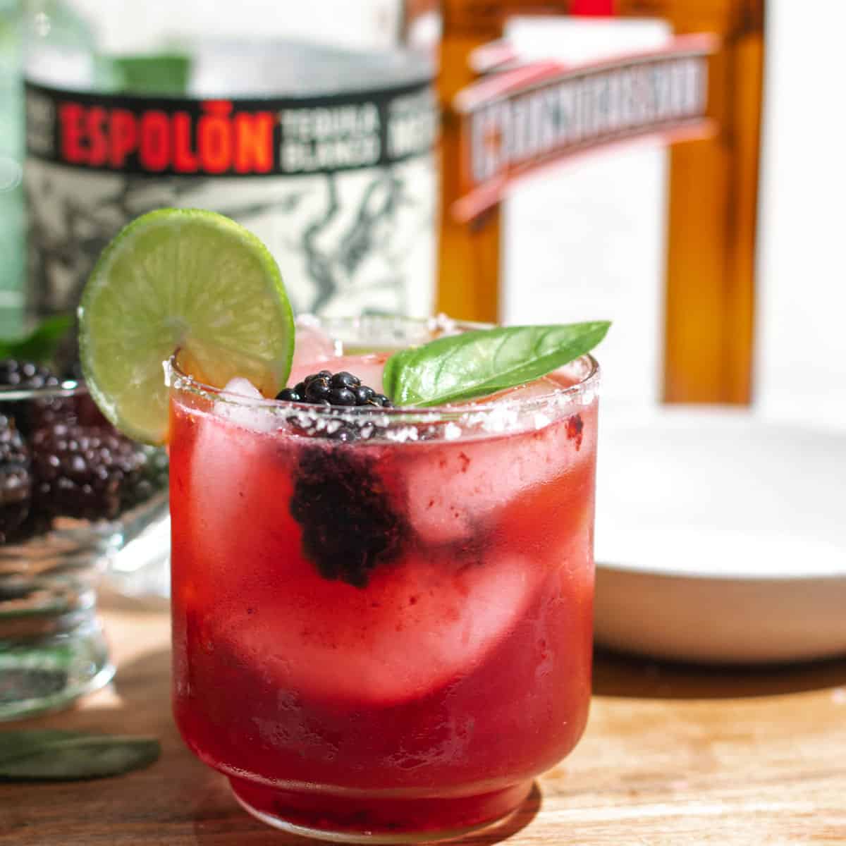 Blackberry basil margarita garnished with blackberries, lime, and basil leaf sitting on a cutting board with a bowl of blackberries, bottle of tequila, and bottle of Cointreau in the background.