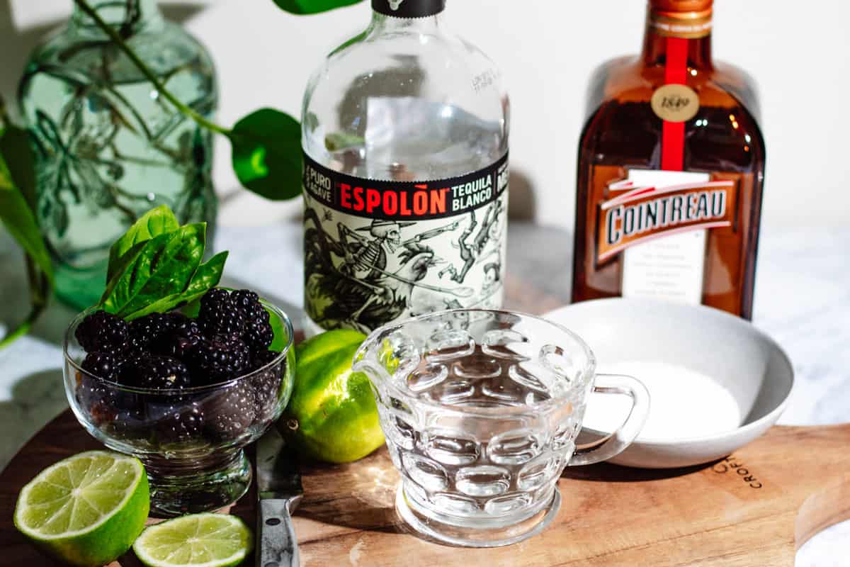 Ingredients for the margaritas on a cutting board including a bowl of blackberries, simple syrup in a glass pitcher, limes, a bottle of tequila, and a bottle of cointreau.