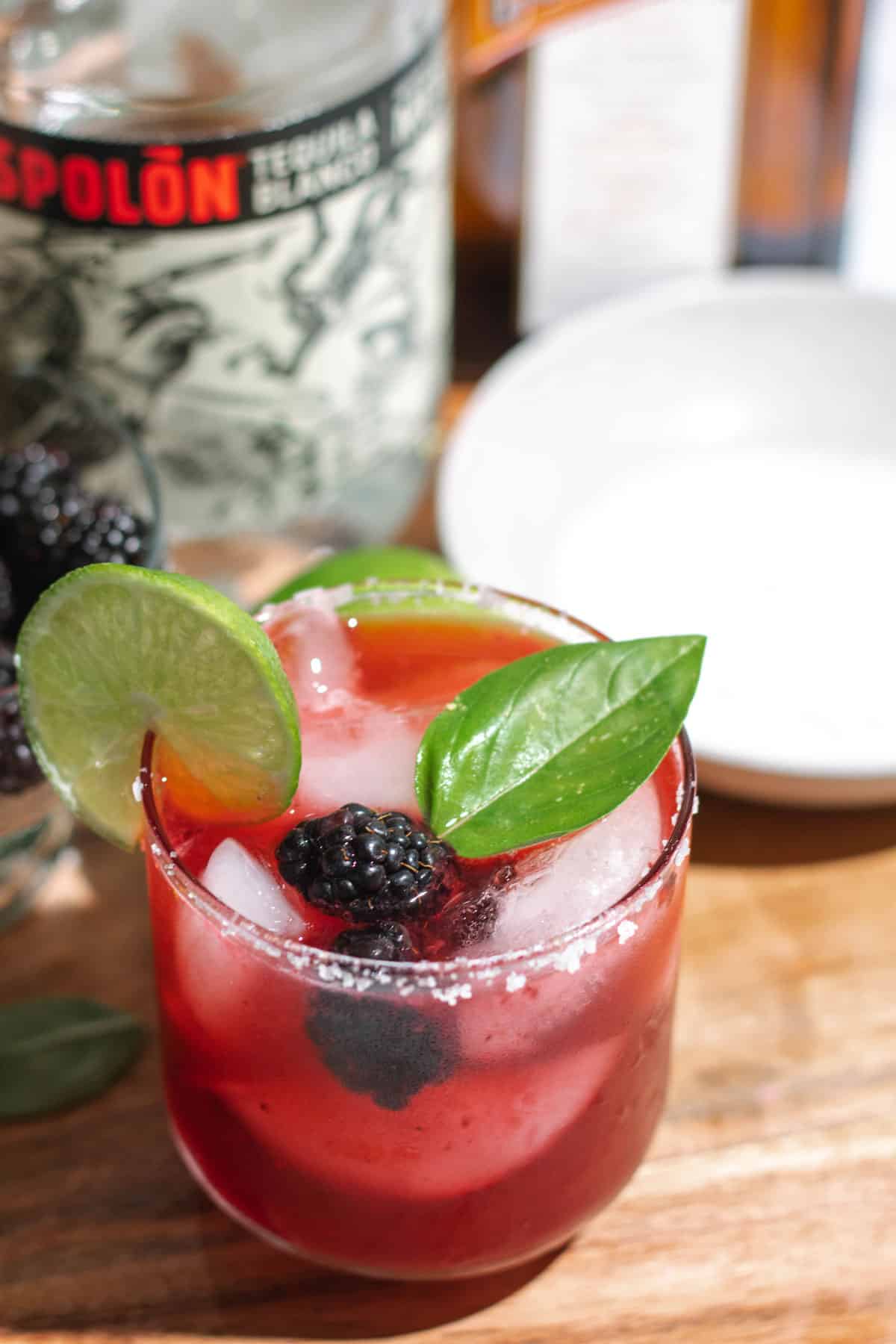 Top of margarita garnished with a basil leaf, blackberries, and a lime wheel on a cutting board with a tray of salt and bottle of tequila in the background.