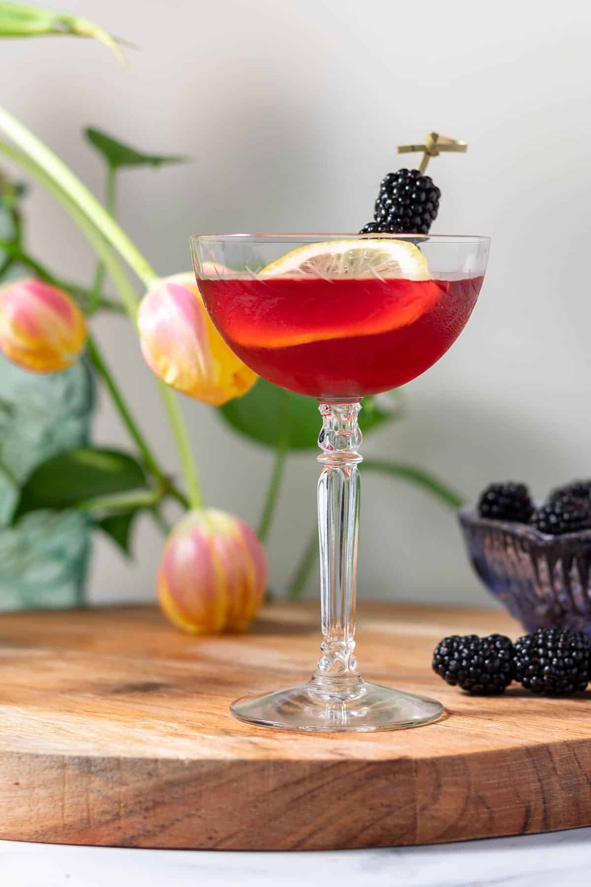 Close up of an empress gin french martini in a coupe glass on a wooden cutting board with a bowl of blackberries and pink tulips in the background.