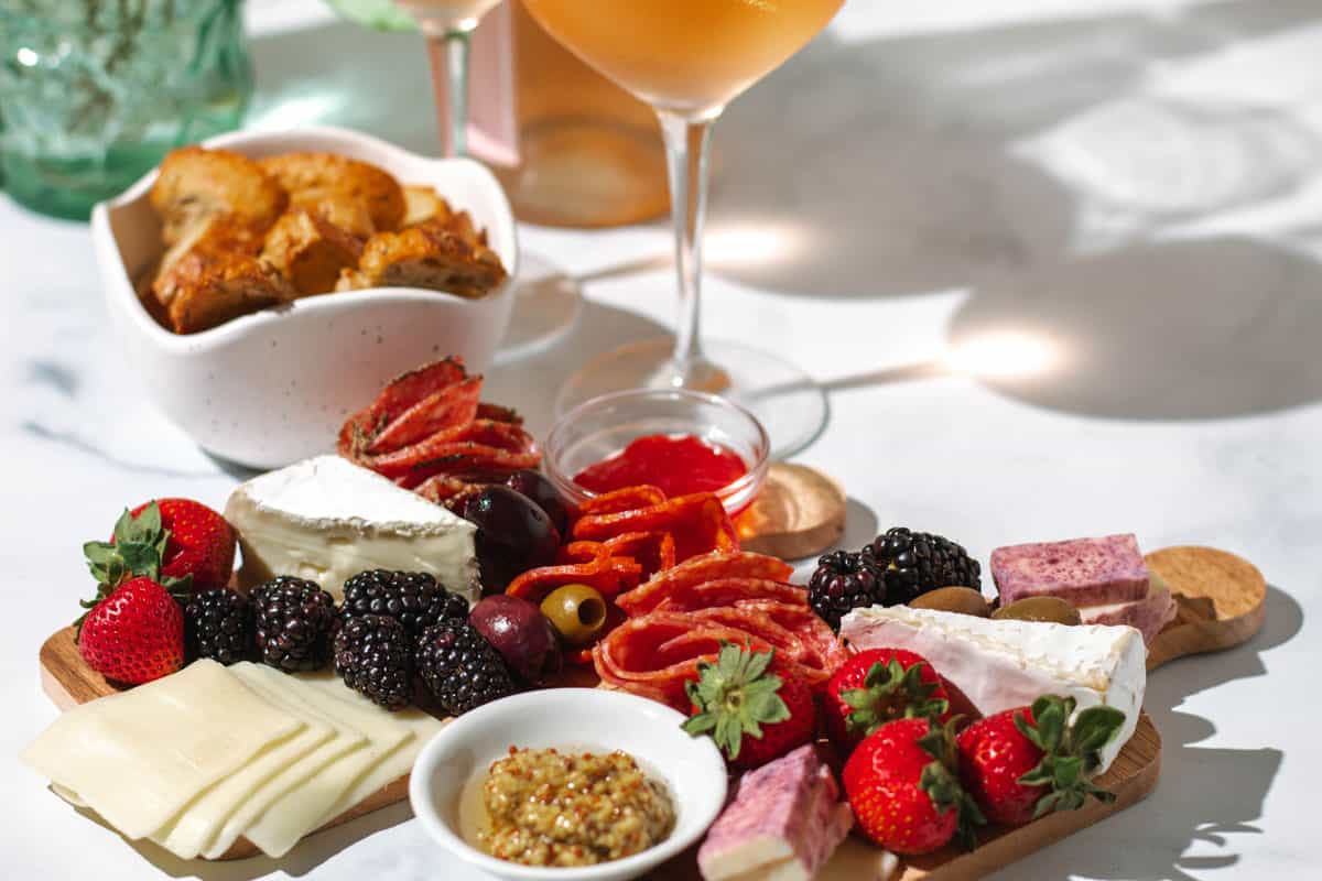 Two mini charcuterie boards with salami, cheddar, brie, mustard, and fruit on a white counter top with 2 glasses of rose wine in the background.