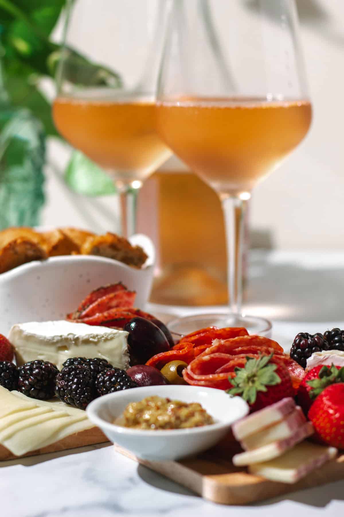 Two mini charcuterie boards with salami, cheddar, brie, mustard, and fruit on a white counter top with 2 glasses of rose wine and bowl of bread in the background.