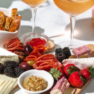 Two glasses of rose wine with two mini charcuterie boards filled with cheeses, meats, and fruit in front and a white bowl of bread on the side.