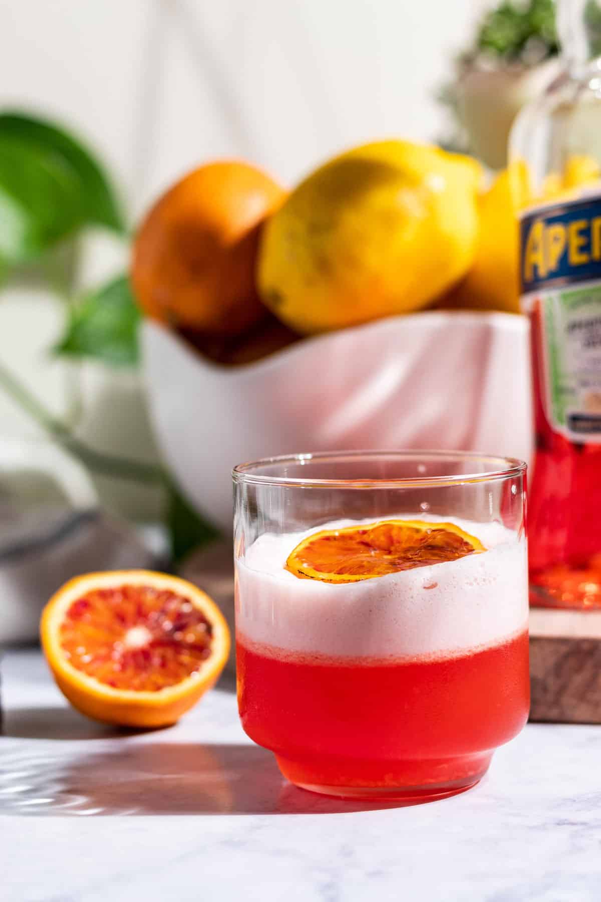 An Aperol sour cocktail with a blood orange half and a bowl of citrus in the background.