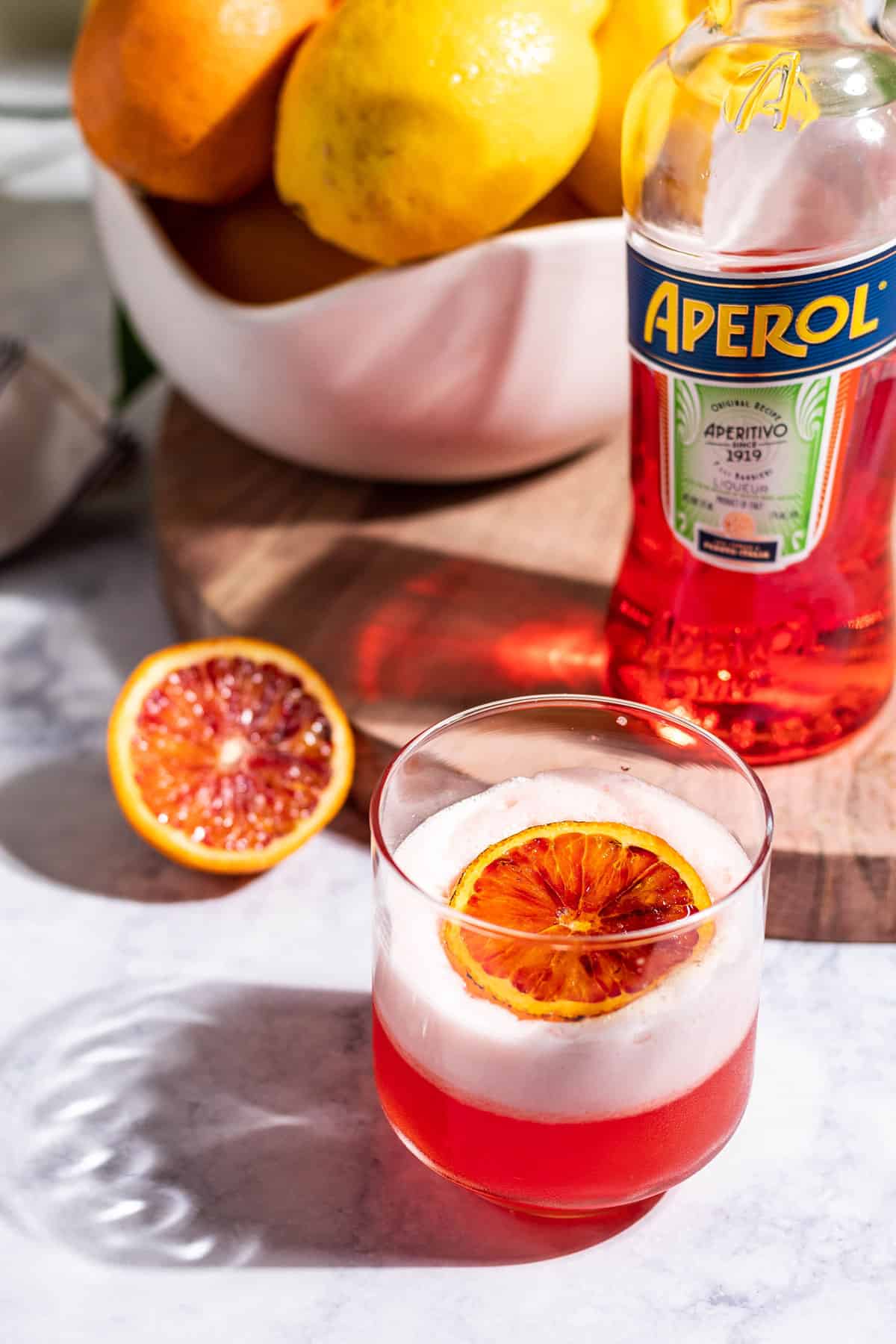 An aperol sour on a white table with a half of blood orange, a bottle of Aperol, and a bowl of citrus in the background on a wood cutting board.