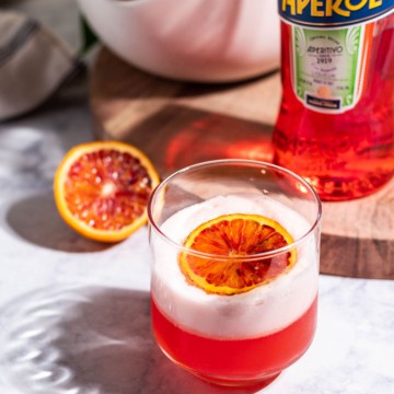 An aperol sour on a white table with a half of blood orange, a bottle of Aperol, and a bowl of citrus in the background on a wood cutting board.