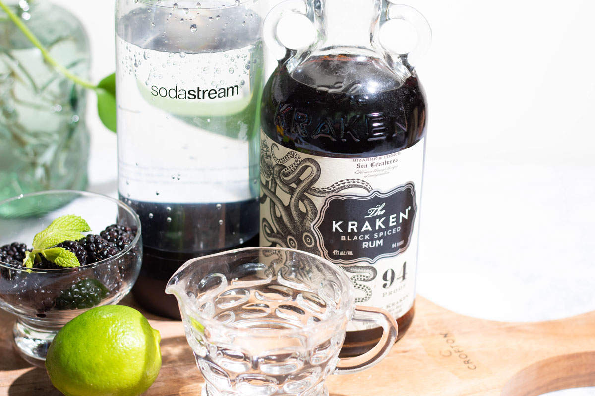 Ingredients for mojitos on a cutting board - including dark rum, seltzer, simple syrup, limes, and blackberries.