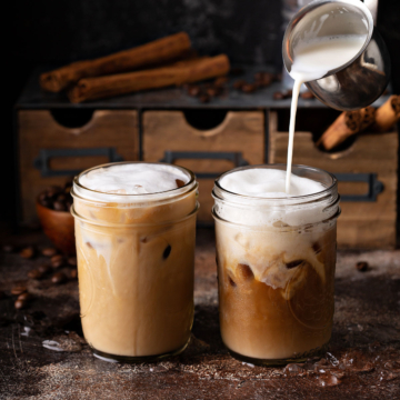 Two iced coffees on a brown, rustic surface with a small metal pitcher pouring in milk.