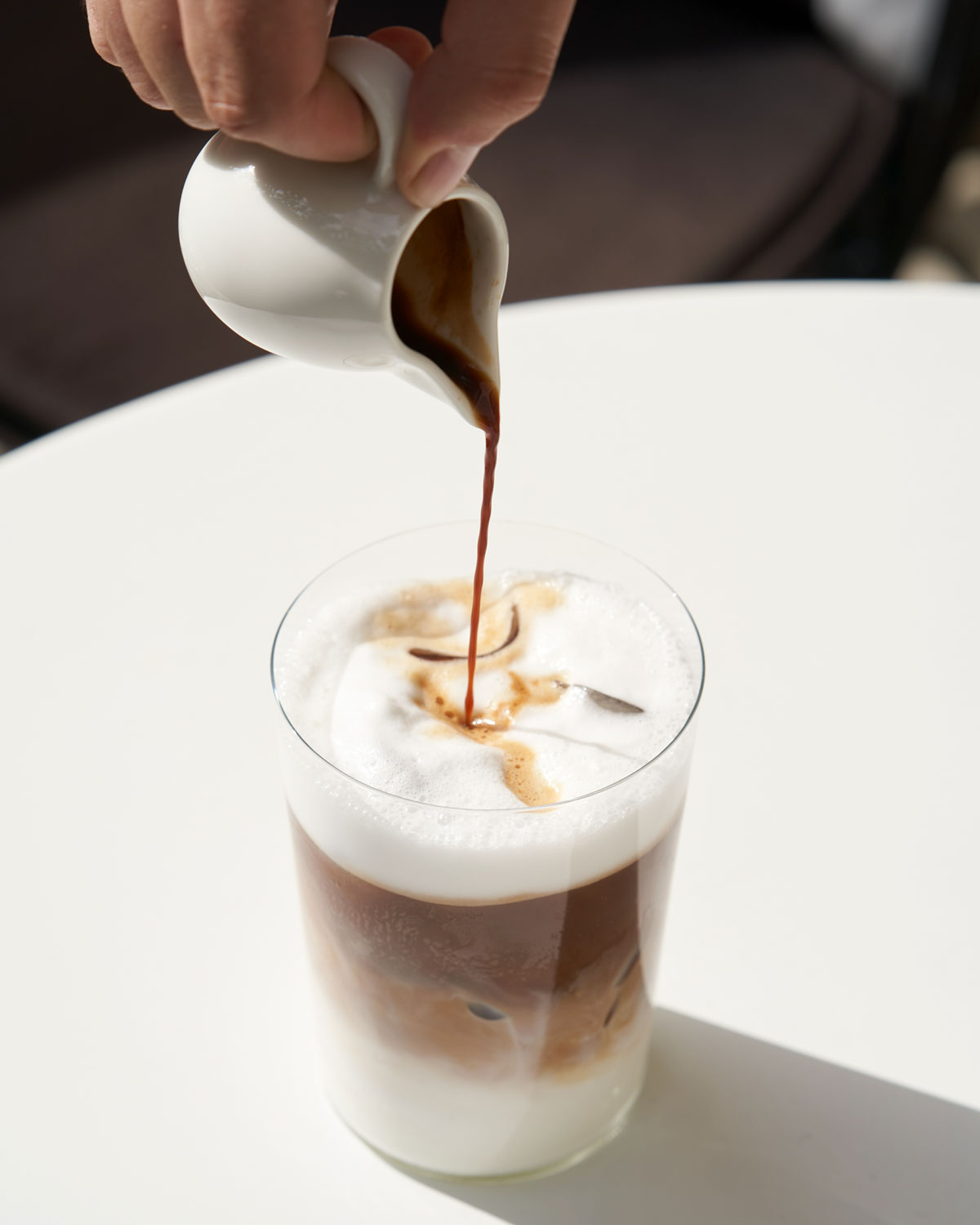 Iced latte on a white table with a small pitcher of syrup being poured on top.