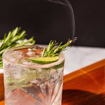 Gin and tonic with a spring of rosemary smoking sitting on a wood cutting board with a bowl of rosemary in the background.