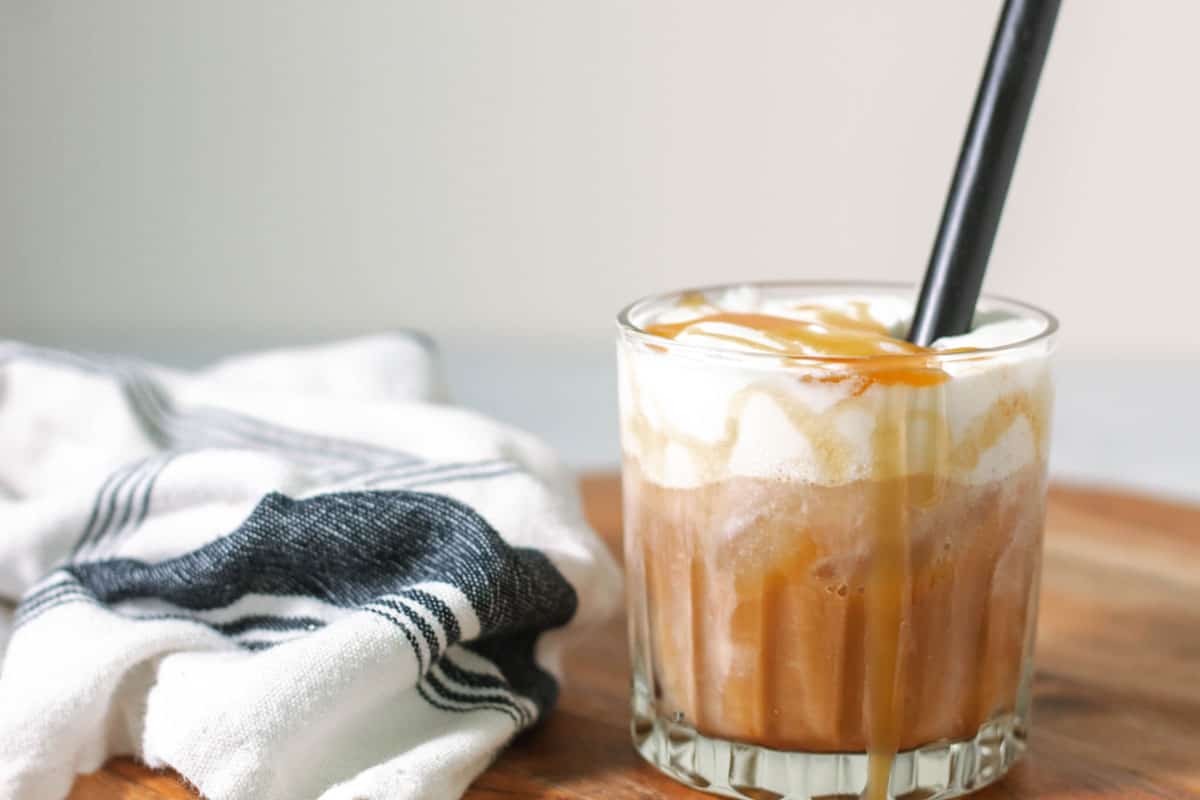 A cup of iced latte on a cutting board with a black straw and caramel rolling down the side of the cup.