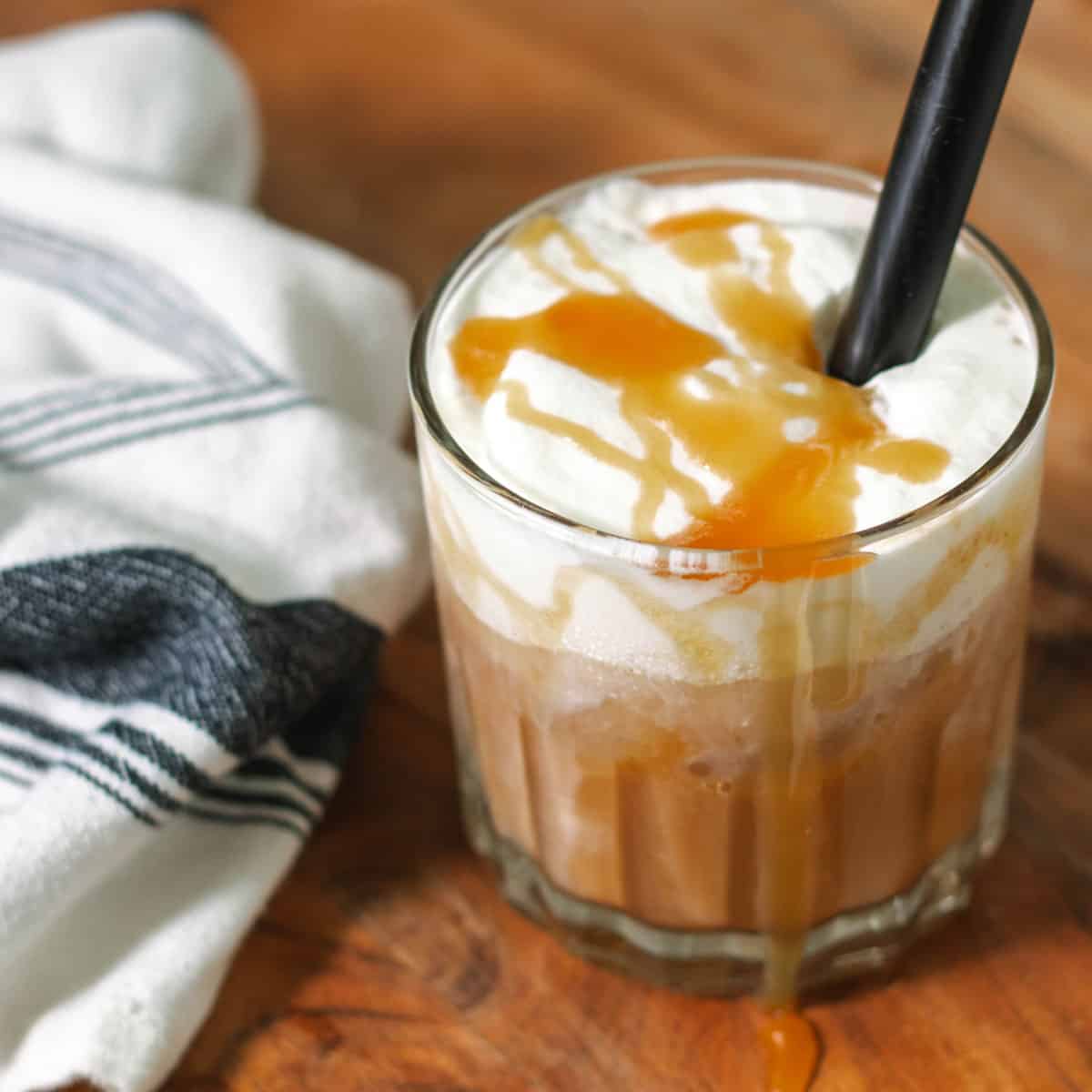 An overview of the iced caramel latte on a cutting board with a black straw and a blue and white napkin on the side.