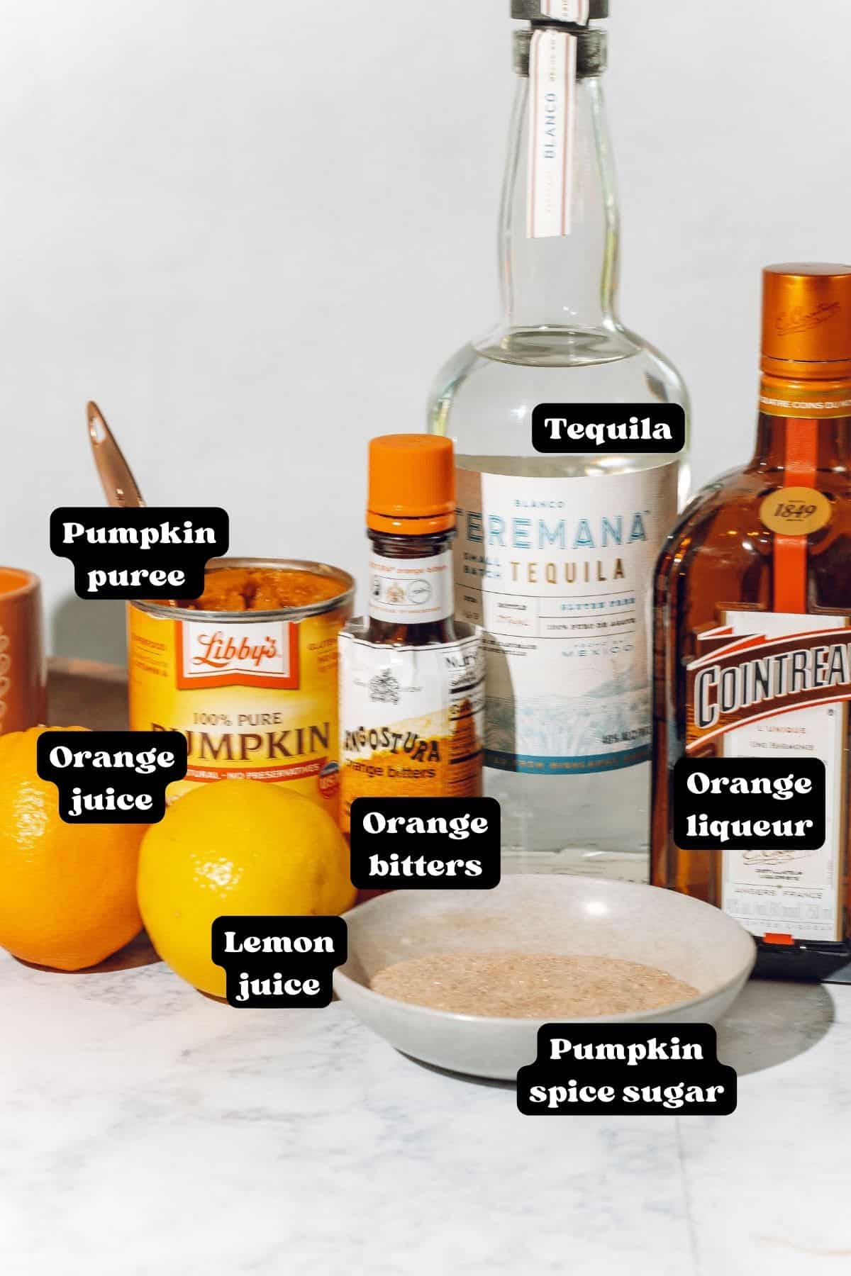 Ingredients for a pumpkin spice margarita on a white surface including tequila, cointreau, pumpkin spice sugar, bitters, pumpkin puree, and citrus fruits.