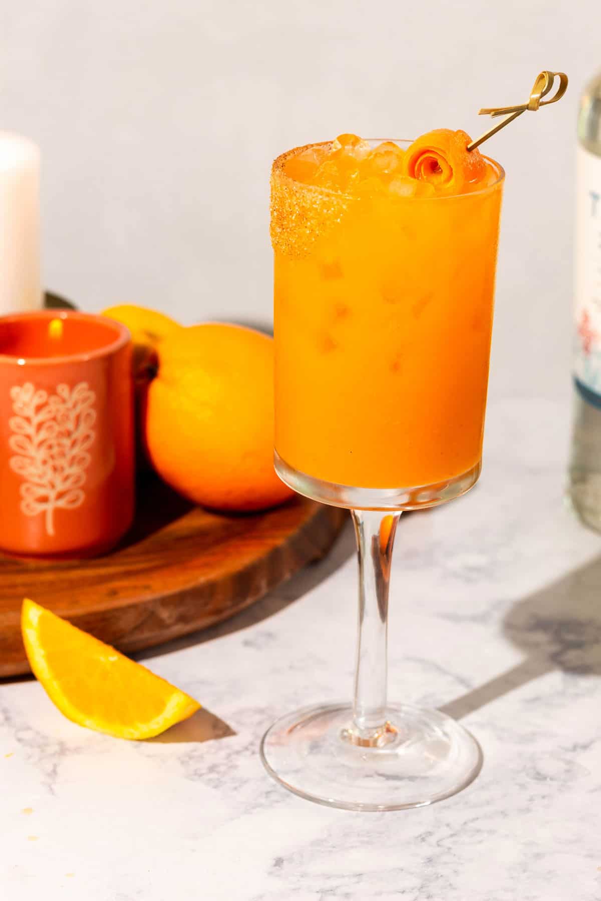 An orange cocktail in a stemmed wine glass with candles, citrus fruit, and tequila in the background.