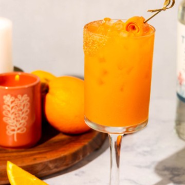 An orange cocktail in a stemmed wine glass with candles, citrus fruit, and tequila in the background.