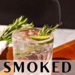 Gin and tonic with a spring of rosemary smoking sitting on a wood cutting board with a bowl of rosemary sprigs in the background. The text overlay reads "smoked gin and tonic".