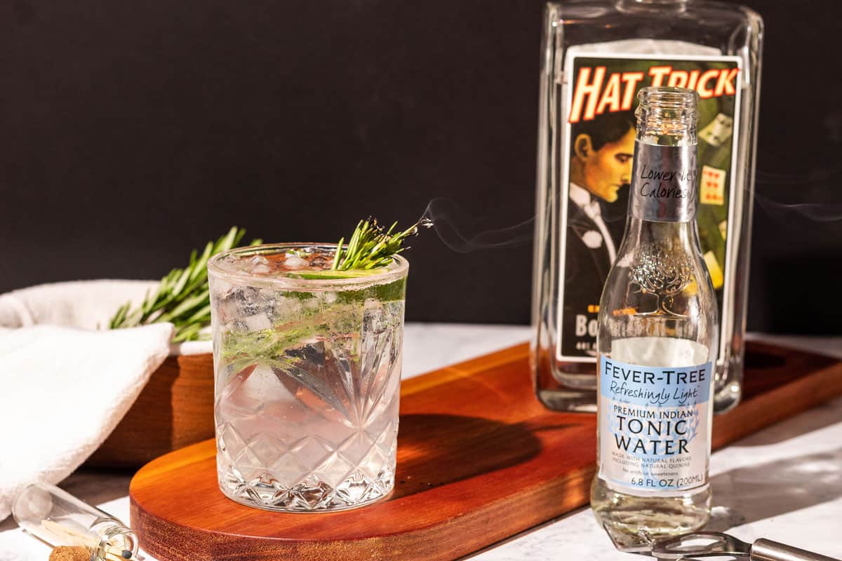 Smoked gin cocktail on a wood cutting board next to a bottle of Hat trick gin and Fevertree tonic water with a black background.