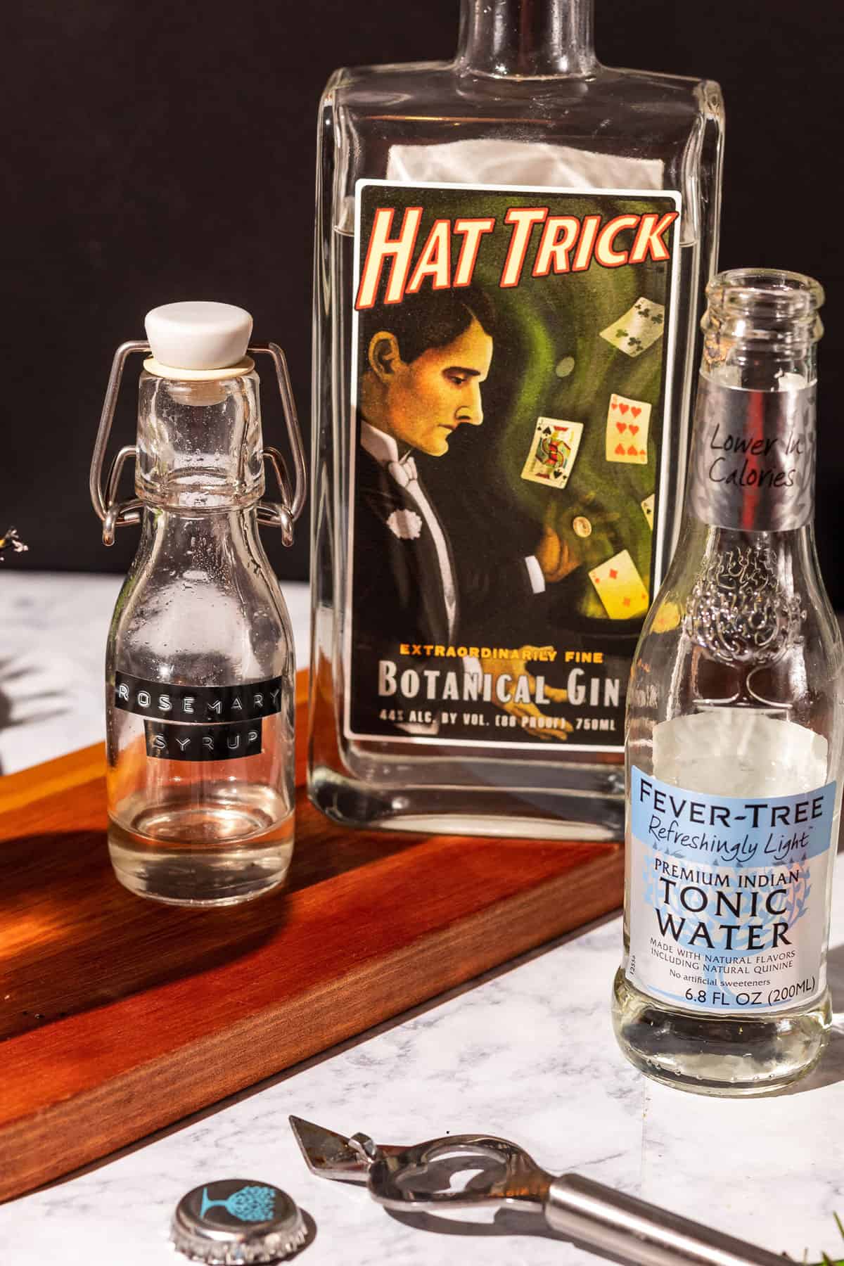 Ingredients for a smoked gin and tonic cocktail - rosemary syrup, gin, and tonic water, on a wood cutting board with a black background.